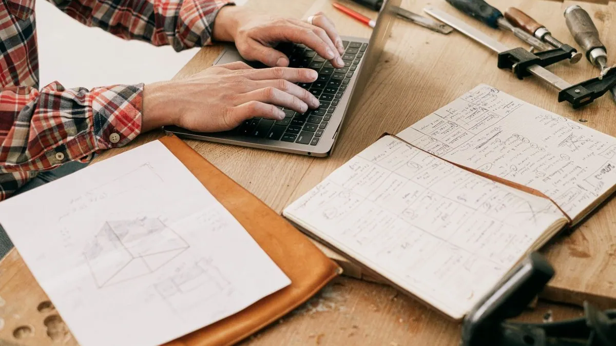 Hands using a laptop beside woodworking tools and project notes